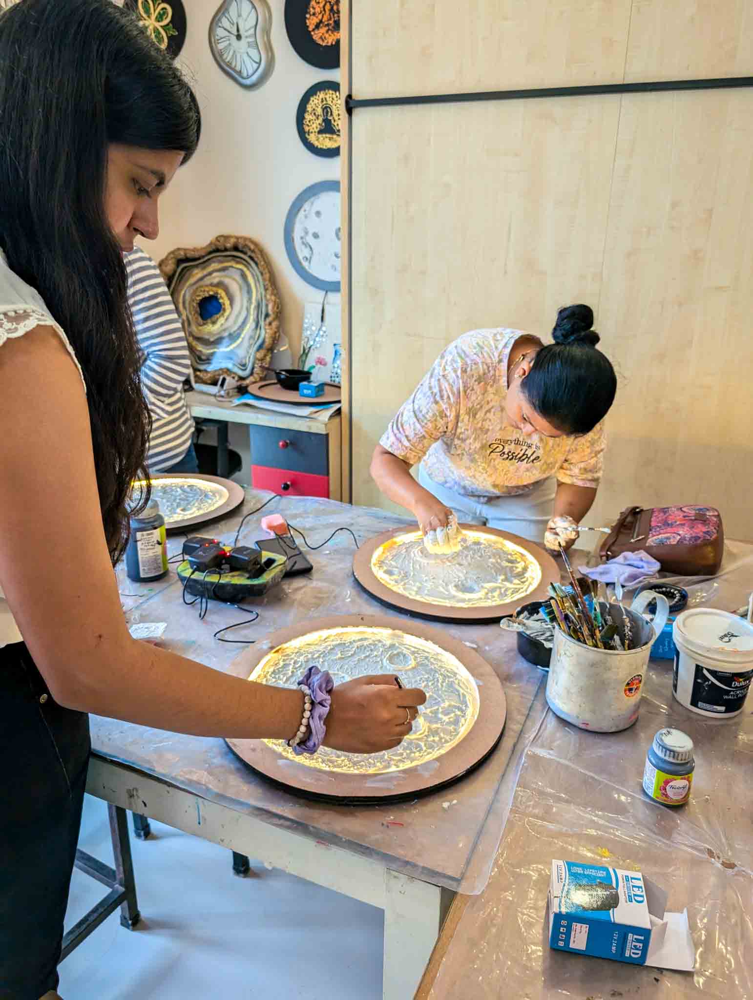 Two women focused on painting and detailing glowing moon lamps on a workshop table.